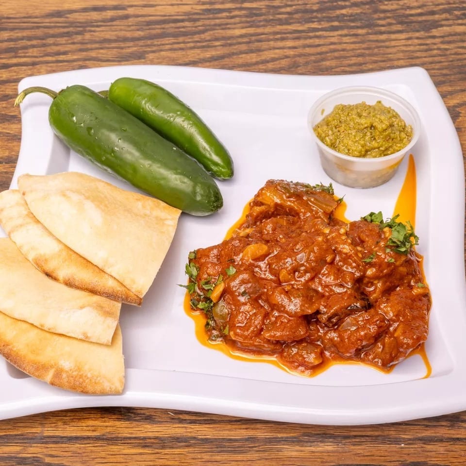 Spicy stew with pita bread, green peppers, and a side of green sauce on a white plate.