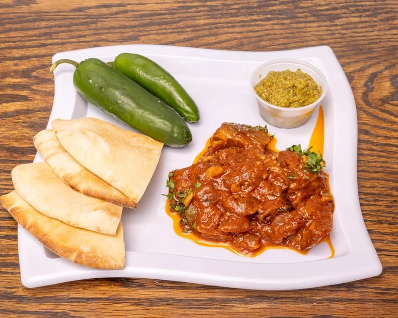 Spicy stew with pita bread, green peppers, and a side of green sauce on a white plate.