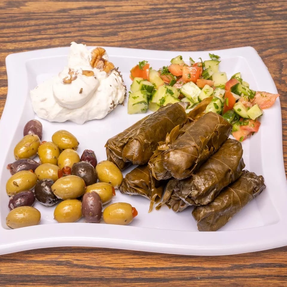 A white plate with dolmades, olives, labneh with walnuts, and a cucumber tomato salad.