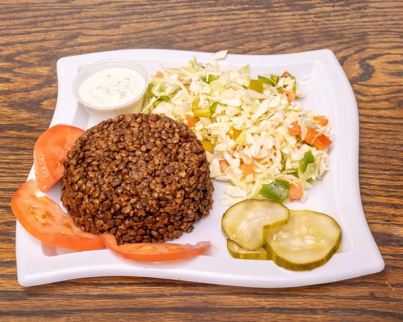 A plate of lentils, coleslaw, tomato slices, pickles, and a side of white sauce.