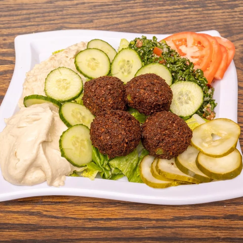 A white plate with falafel, hummus, tabbouleh, sliced cucumbers, tomatoes, and pickles.
