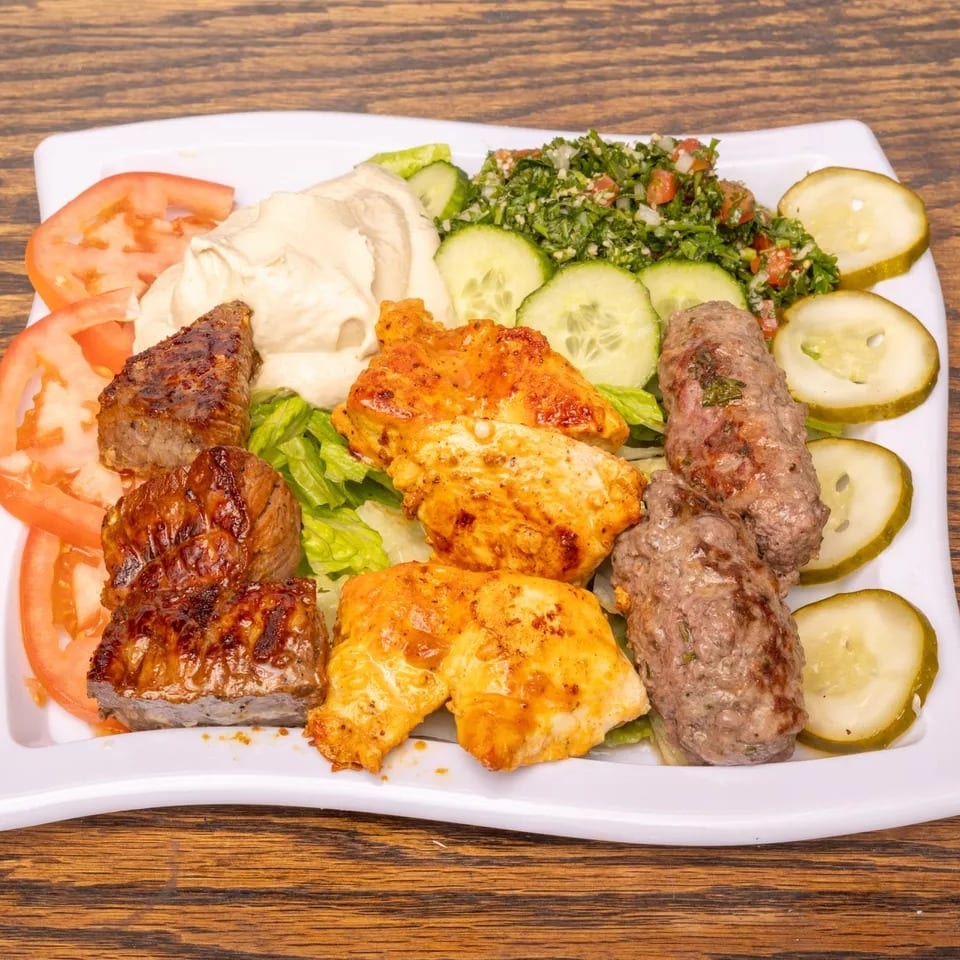 Grilled meat platter with chicken, beef, and kofta, served with hummus, tabbouleh, tomatoes, and pickles.