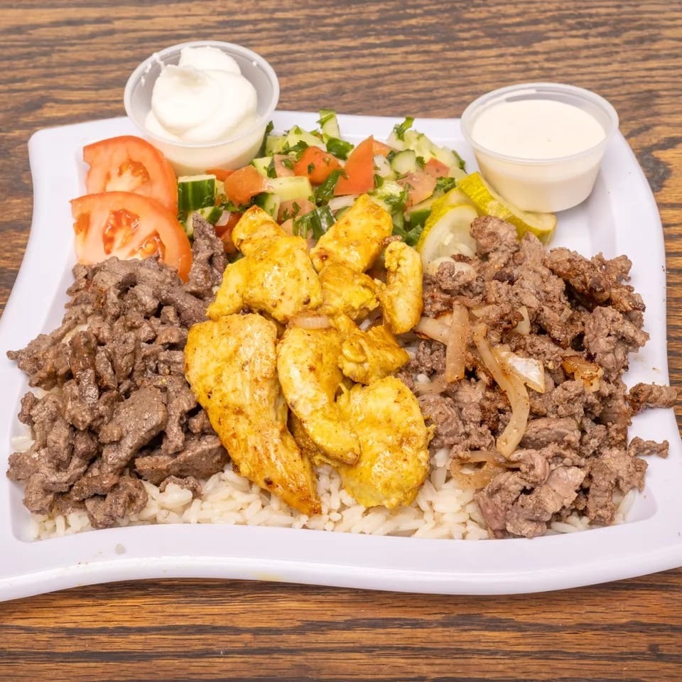 A platter of rice topped with chicken and beef shawarma, salad, tomatoes, pickles, and two sauces.