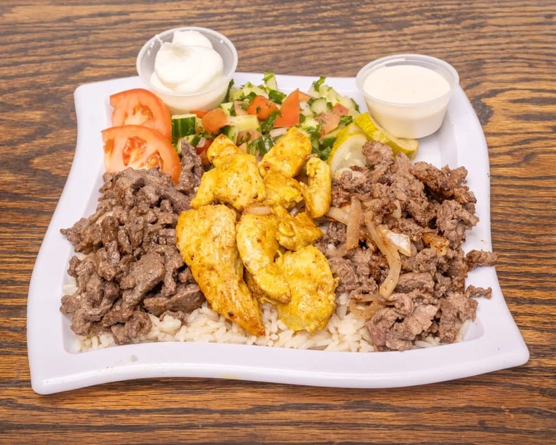 A platter of rice topped with chicken and beef shawarma, salad, tomatoes, pickles, and two sauces.
