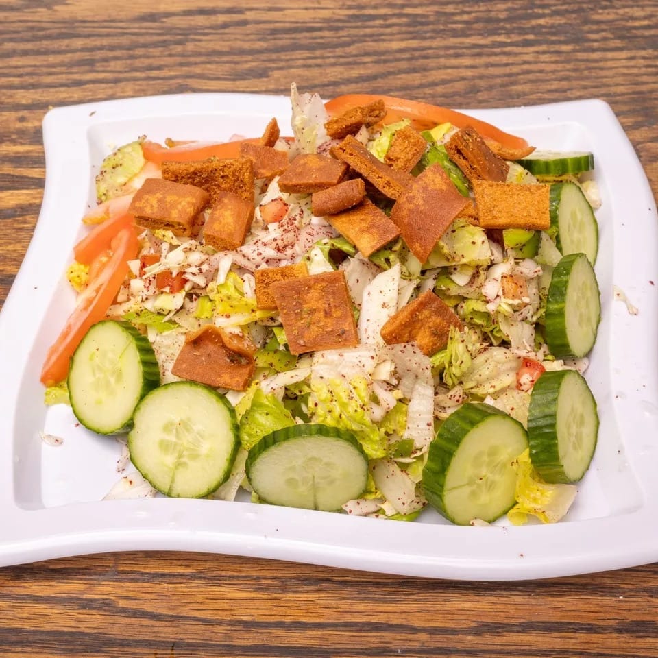 A white rectangular plate of fattoush salad with lettuce, cucumbers, tomatoes, and toasted pita bread.