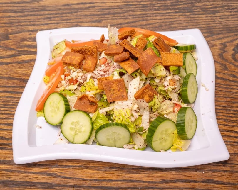 A white rectangular plate of fattoush salad with lettuce, cucumbers, tomatoes, and toasted pita bread.